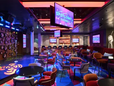 Karaoke-Bar 'Spotlight Karaoke' auf der Icon of the Seas. Bühne mit Mikrofon und Lautsprecherwand. Bunte Sitzmöbel. Beleuchtete Bar. Farbiges Licht.