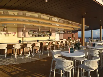 Open-Air-Bar „Hideaway“ auf der Icon of the Seas mit langem Tresen, weißen Hockern und Sitzgruppen. Regale mit Flaschen. Meer im Hintergrund.