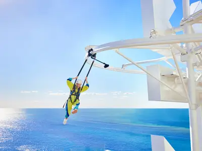 Eine Person in Schutzkleidung ist bei einer Outdoor-Attraktion der Icon of the Seas hoch über dem Ozean zu sehen. Sie schwebt an Gurten und Stangen, mit blauem Himmel und glitzerndem Meer im Hintergrund.