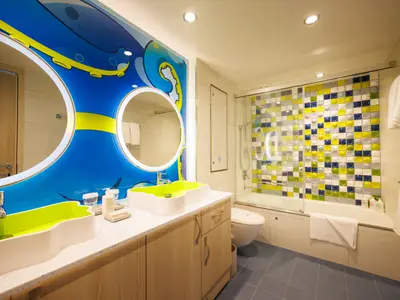 Modernes Badezimmer an Bord der Icon of the Seas. Zwei beleuchtete Spiegel, grüne Waschbecken, Dusche über Badewanne mit bunten Fliesenwand und blauer Wanddekor.