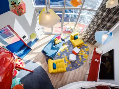 Draufsicht auf eine farbenfrohe Familiensuite der Icon of the Seas. Ein Wohnbereich mit blauer Couch, gelben Sitzen, einem geometrischen Teppich und einer roten Rutsche. Große Fenster bieten Meerblick.