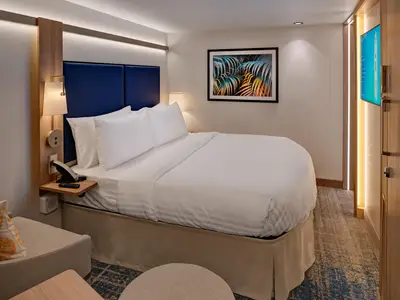 Eine moderne Kreuzfahrtkabine mit einem gemachten Doppelbett in Weiß. Links ein Holznachttisch mit Telefon und Lampe. An der Wand ein farbenfrohes Kunstbild. Der Teppichboden ist gemustert.