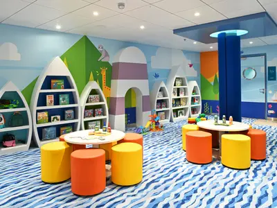 Heller Spielbereich auf der Icon of the Seas. Bootsregale mit Büchern, bunte Tische mit Hockern, wellenförmiger Teppich und maritime Wandmalerei mit Tieren.