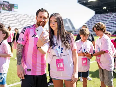 Fussballspieler Lionel Messi macht ein lächelndes Selfie mit einem jungen Mädchen auf einem Spielfeld. Beide sind pink gekleidet, weitere Kinder in pinken Shirts im Hintergrund.