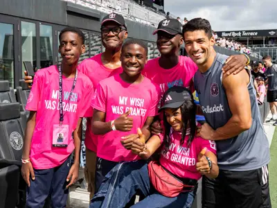 Eine lächelnde Gruppe von Jugendlichen und Erwachsenen in pinken T-Shirts, die neben Fussballstar Luis Suárez posieren, im Inter Miami CF Stadion. Die T-Shirts tragen die Aufschrift 'MY WISH WAS GRANTED'. Ein Jugendlicher trägt eine VIP-Karte um den Hals. Im Hintergrund sind Stadiontribünen zu sehen.