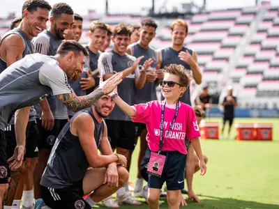 Lachender Junge in pinkem Shirt gibt Fussballstar Lionel Messi auf einem grünen Feld ein High-Five. Spieler und Stadion im Hintergrund. Der Junge trägt VIP-Band.