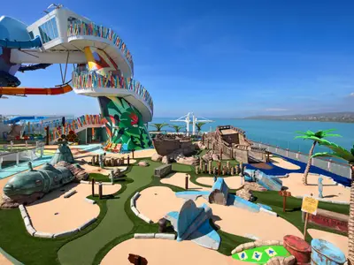 Ansicht des thematischen Mini-Golf-Platzes an Bord der Icon of the Seas. Im Vordergrund Schiffswrack-Elemente, im Hintergrund bunte Wasserrutschen, das Meer und eine Küste.