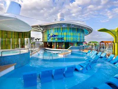 Der belebte Poolbereich der Icon of the Seas zeigt die Swim & Tonic Bar, blau untergetauchte Liegen, einen Whirlpool und bunte Strukturen unter dem bewölkten Himmel.