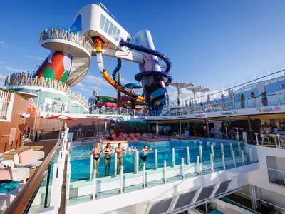 Das belebte Oberdeck der Icon of the Seas zeigt mehrere Pools mit Glaswänden, farbenfrohe Wasserrutschen, Sonnenliegen und Bars unter blauem Himmel. Mehrere Personen entspannen und baden.