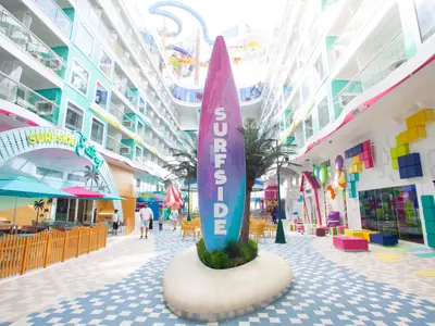 Das Surfside-Viertel der Icon of the Seas. Zentrale Surfbrett-Skulptur, bunte Balkone, Essbereiche und Wasserrutschen im Hintergrund. Lebhaft und farbenfroh.