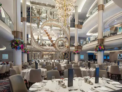 Das Hauptrestaurant der Icon of the Seas zeigt gedeckte Tische, eine mehrstöckige goldene Kunstinstallation und bunte Blumendekorationen.