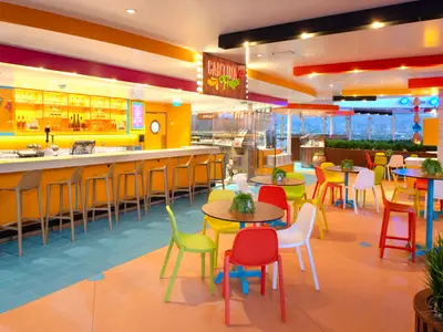 Cantina Fresca auf der Icon of the Seas. Der farbenfrohe Raum zeigt eine lange Bar mit Hockern, Tische mit bunten Stühlen und den Blick auf eine Stadt bei Nacht.