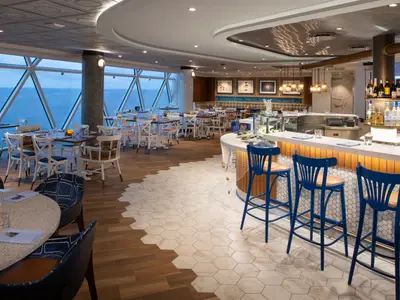 Restaurant auf der Icon of the Seas mit Meerblick durch große Fenster. Viele Tische, eine Bar mit blauen Hockern, Boden aus Holz und Hexagonfliesen.