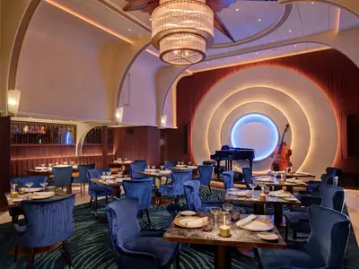 Restaurant auf der Icon of the Seas mit blauen Samtstühlen, Tischen, Bühne mit Flügel und Kontrabass. Art-déco-Wanddesign mit Leuchtkreisen.