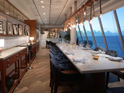 Das elegante Restaurant der Icon of the Seas zeigt eine lange, gedeckte Tafel mit Meerblick. Auffällig sind die Seil-Pendelleuchten und die maritim gestalteten Wände mit Bildern und Spiegeln.