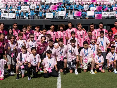 Eine Gruppe von Fussballspielern in pinkfarbenen Trikots posiert mit lachenden Kindern auf einem sonnigen Rasenplatz. Im Hintergrund jubelnde Fans mit "Make-A-Wish"-Schildern auf der Tribüne.