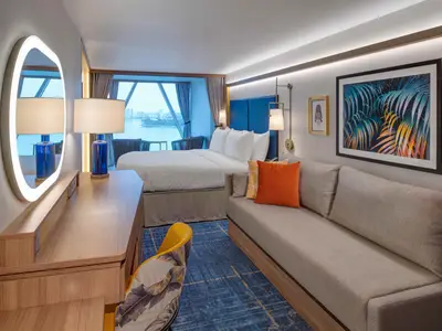 Kabine der Icon of the Seas mit Doppelbett, Sofa, Schreibtisch. Große Fensterfront zeigt Meer und Stadt. Modernes Design mit blauen und orangen Details.