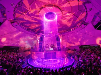 Zwei Akrobaten schweben über einer Bühne mit einem beleuchteten Wasserbecken und Fontänen. Ein riesiger Discoball oben sprüht Wasser, während Lichtstrahlen die Szene in Blau- und Violetttönen illuminieren. Modernes Theater mit Zuschauersitzen seitlich.