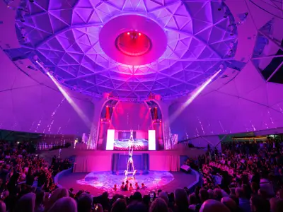 Weitwinkelaufnahme einer lebhaften Akrobatik-Show in einem großen, kuppelartigen Saal. Artisten vollführen Figuren im Wasserbecken und in der Luft unter lila Beleuchtung, während ein großes Publikum zuschaut.