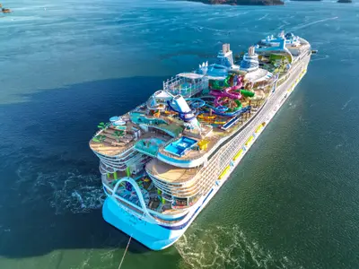 Luftaufnahme der Icon of the Seas. Das Schiff liegt ruhig im blauen Wasser. Bunte Wasserrutschen, Pools und Decks mit Grünflächen sind sichtbar. Der Schiffsname ist am Heck erkennbar.