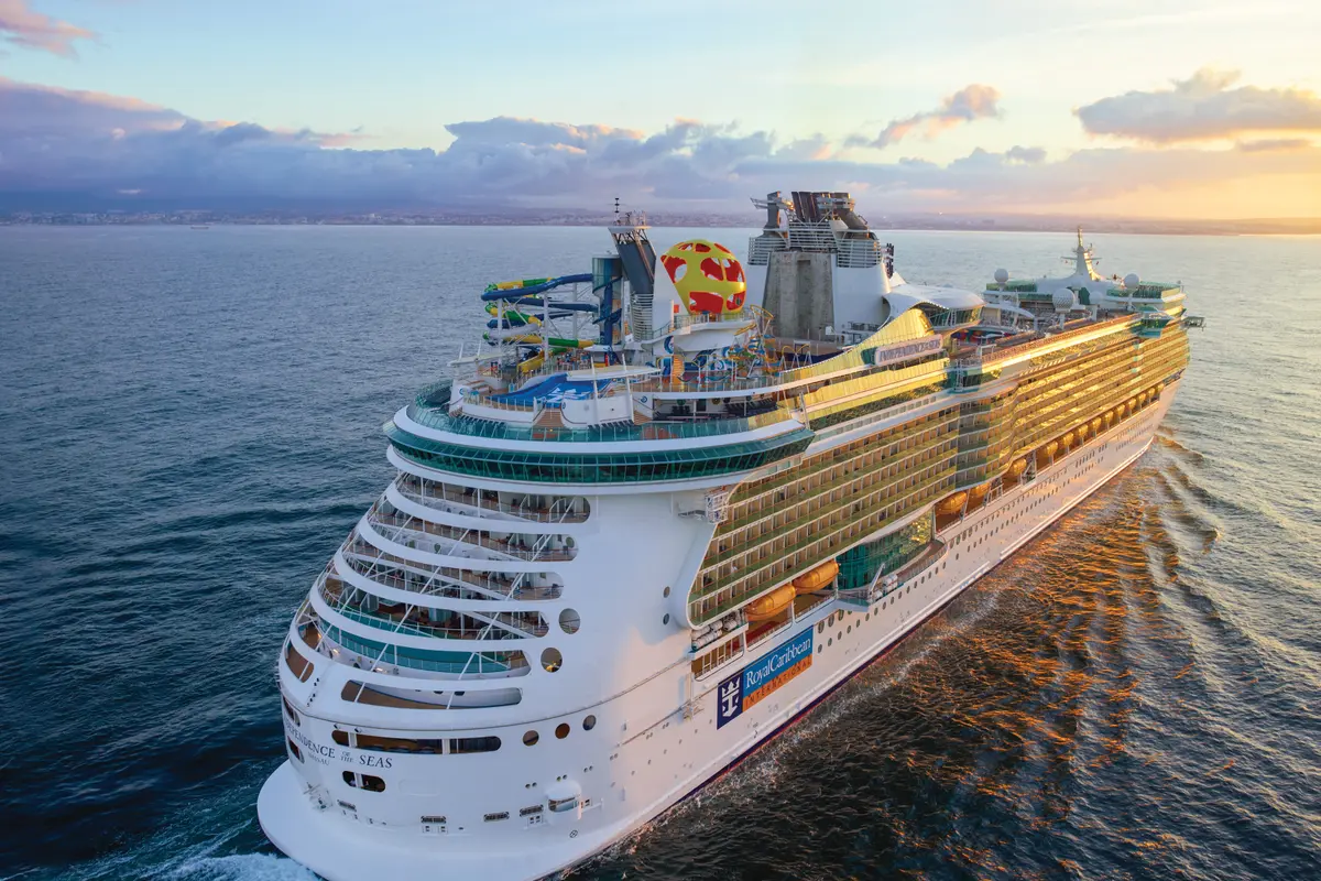 Blick aus der Vogelperspektive auf die Independence of the Seas, ein großes Schiff von Royal Caribbean. Die Sonne geht unter und taucht das Schiff, das Meer und den Himmel in warmes, goldenes Licht. An Deck sind bunte Wasserrutschen und Freizeiteinrichtungen sichtbar, während das Schiff eine leichte Heckwelle zieht.