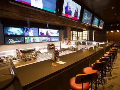 Innenansicht der 'Playmakers' Sports Bar auf der Independence of the Seas. Lange Theke mit roten Hockern. Bildschirme zeigen Sport und 'Playmakers'-Logo.