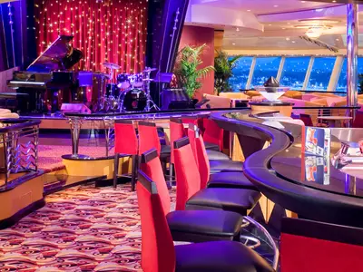 Innenansicht der Independence of the Seas: Eine Bar mit Bühne, Klavier, Schlagzeug, roten Vorhängen und Barhockern. Nächtlicher Stadtblick durch die Fenster.