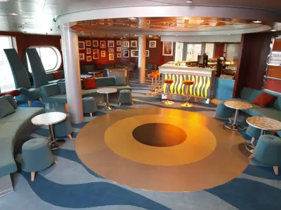 Lounge auf der Independence of the Seas mit beleuchteter Bar, türkisfarbenen Sitzgelegenheiten, Tischen und einem Teppich mit Wellen- und Kreisdesign. Fenster sind sichtbar.
