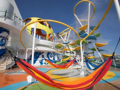 Sonniges Deck der Independence of the Seas mit bunten Hängematten, einer Kletterstruktur aus Netzen und farbigen Blattformen. Blauer Himmel und Wellen-Mural.