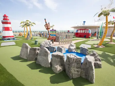 Blick auf die Mini-Golfanlage an Deck der Independence of the Seas. Zu sehen sind ein rot-weißer Leuchtturm, Palmen, ein Wegweiser und ein roter Bus. Im Vordergrund Felsen und ein Wasserlauf.