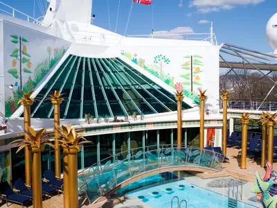 Ein sonniges Deck der Independence of the Seas zeigt einen Pool mit einer geschwungenen Brücke, goldene Säulen und eine große, schräge Glaskuppel. Die umliegenden Wände sind mit tropischen Pflanzen bemalt.