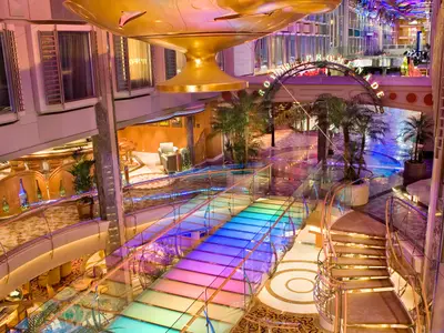 Das Bild zeigt die mehrstöckige Royale Promenade der Independence of the Seas. Eine bunte, beleuchtete Glasbrücke führt über die Halle, umgeben von Sitzbereichen, Geschäften und Palmen. Ein großes goldenes Objekt hängt von der Decke.