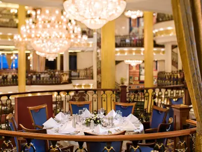 Gedeckter Tisch mit blauen Stühlen auf einem Balkon im Inneren der Independence of the Seas. Im Hintergrund das helle, mehrstöckige Atrium mit Kronleuchtern.