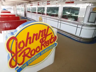 Das Johnny Rockets Diner auf der Independence of the Seas. Rote Sitzbänke, Tische und ein Diner-Tresen sind zu sehen. Vorne ein großes Johnny Rockets Logo.