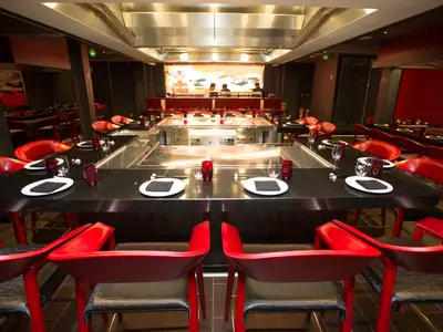 Modernes Teppanyaki-Restaurant der Independence of the Seas. Rote Stühle und schwarze Tische umgeben Kochstationen. Köche arbeiten im Hintergrund an der Sushi-Bar.