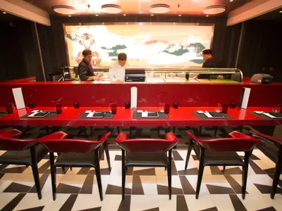 Das moderne Sushi-Restaurant an Bord der Independence of the Seas, mit drei Köchen hinter einer roten Theke und roten Stühlen im Vordergrund.