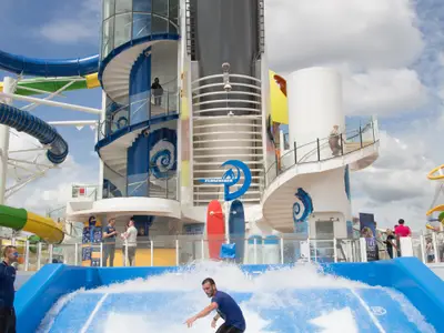 Ein Surfer auf dem FlowRider-Simulator der Independence of the Seas. Im Hintergrund sind farbige Wasserrutschen, Wendeltreppen und weitere Passagiere auf dem Deck unter teils bewölktem Himmel zu sehen.