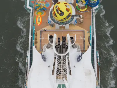 Luftbild des Oberdecks der Independence of the Seas. Man sieht bunte Freizeitanlagen, darunter ein gelbes SkyPad, einen Basketballplatz, die großen Schornsteine und das schäumende Meer entlang des Schiffs.