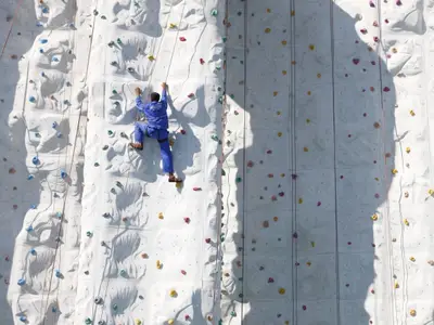 Eine Person in blauer Kleidung klettert an einer weißen Kletterwand mit bunten Griffen an Bord der Independence of the Seas. Mehrere Seile sichern den Aufstieg unter hellem Sonnenlicht.