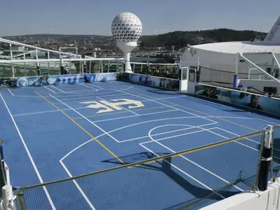Sportplatz auf dem Deck der Independence of the Seas. Blauer Platz, Basketballkörbe, Logo. Stadt, Hügel und große Golfball-Struktur hinten.
