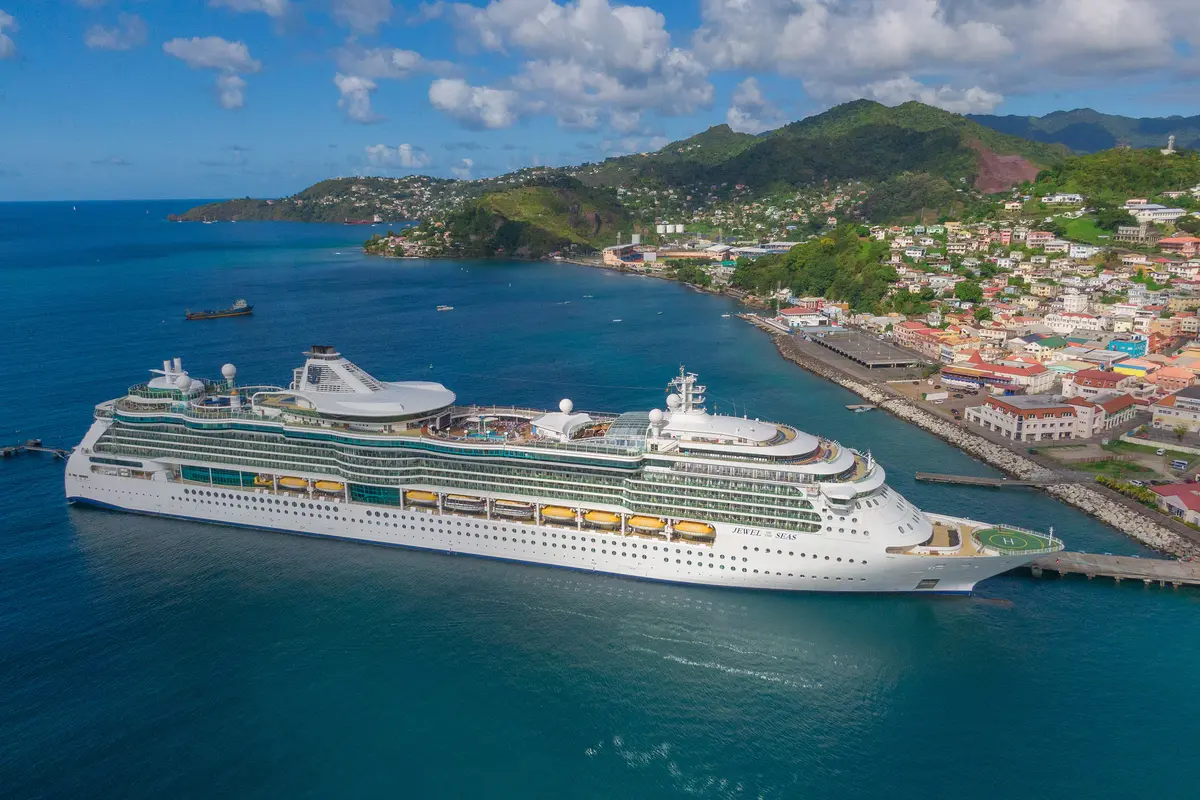 Vogelperspektive der weißen Jewel of the Seas im blauen Wasser eines Hafens. Im Hintergrund eine bunte Küstenstadt und grüne Berge unter blauem Himmel mit Wolken.
