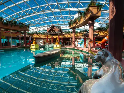 Der Innenpoolbereich der Jewel of the Seas unter einem Glasdach. Das türkisfarbene Wasser spiegelt die Decke. Säulen, Pflanzen und eine Tierstatue sind sichtbar.