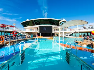 Das sonnige Pooldeck der Jewel of the Seas mit mehreren blau schimmernden Pools. Liegestühle, eine große LED-Leinwand und ein Rettungsschwimmer sind sichtbar. Blauer Himmel über dem Schiff.