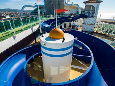 Blick auf das Deck der Jewel of the Seas mit gewundenen blauen Wasserrutschen um einen weiß-blau gestreiften Zylinder mit goldener Kuppel. Im Hintergrund sind Land und Meer zu sehen.