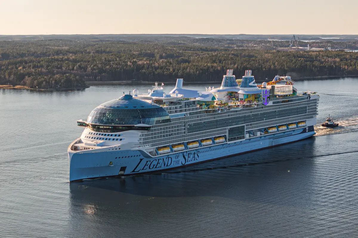 Die große, blaue Legend of the Seas durchquert Gewässer, begleitet von einem Schlepper am Heck. Im Hintergrund sind bewaldete Ufer und der Himmel zu sehen. Die Sonne scheint hell auf das Schiff.