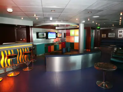 Innenansicht eines farbenfrohen Freizeitbereichs an Bord der Liberty of the Seas. Die Bar, eine Spielecke mit Bildschirmen (Wii Resort) und Sitzgruppen sind sichtbar.