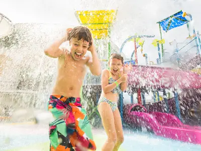 Zwei lachende Kinder spielen im Splashaway Bay Wasserpark der Liberty of the Seas. Sie werden von Wasser fontänen nass gespritzt. Bunte Wasserrutschen sind im Hintergrund sichtbar.