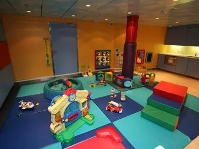Bunter Kinderspielbereich auf der Liberty of the Seas. Der Raum zeigt Spielmatten, diverse Spielzeuge, darunter ein Spielhaus und Bausteine, sowie eine Küchenzeile mit blauen Schränken.