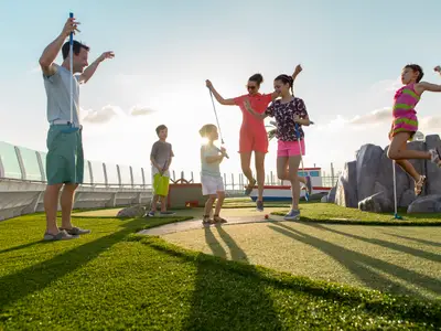 Familie spielt Minigolf auf dem Außendeck der Liberty of the Seas. Erwachsene und Kinder springen vor Freude auf Kunstrasen. Sonnenschein und lange Schatten prägen die Szene.