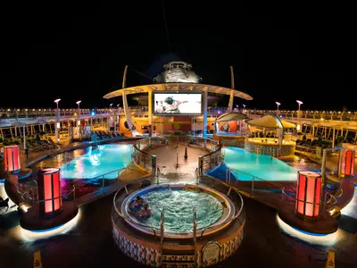 Nachtansicht des beleuchteten Pooldecks der Liberty of the Seas von Royal Caribbean International mit mehreren Pools, Whirlpools, Liegen und einer Großleinwand unter dem Nachthimmel.